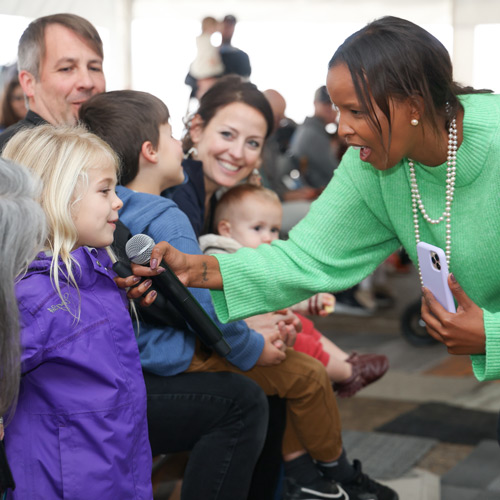 Smiling woman in a green sweater holding a microphone to a young blonde girl in a purple jacket, with a seated audience watching.