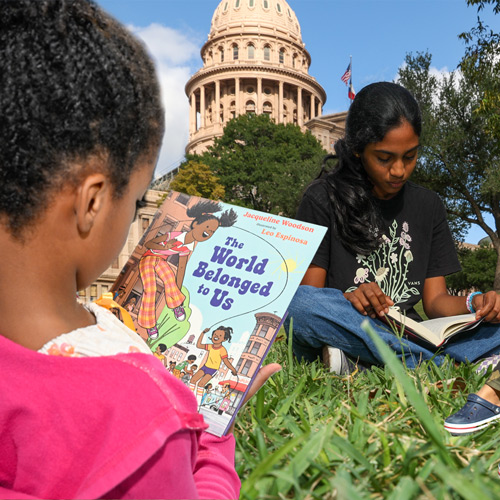 Two girls sit on grass reading books outdoors with the Texas Capitol building in the background. One is reading "The World Belonged to Us" by Jacqueline Woodson and Leo Espinosa.